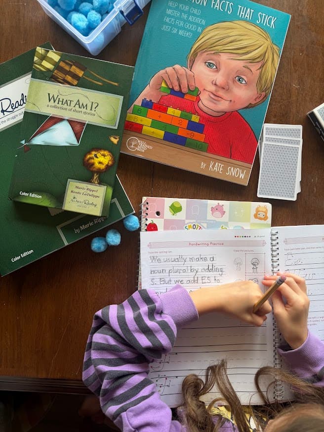 Child writing in a handwriting workbook at a homeschool table with reading and math books